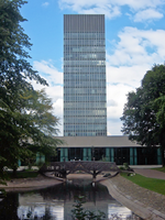 Blue/Grey skyscraper with a glass curtain wall. In the foreground is park pond with a small wooden bridge and trees at either side. In between the Tower and the pond is a modern 2 storey building