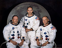 Three astronauts in spacesuits without helmets sitting in front of a large photo of the moon.