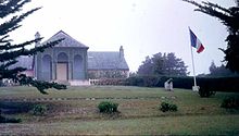 Photo of a front garden and large brown building. French flag on a flagpole next to a small cannon.