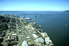 A long bridge crosses a huge river flowing into a vast body of water under a blue sky. The bridge begins in a settlement with streets, buildings, and wharfs along the river and extends out of sight toward a low hill on the far shore. The first part of the bridge has a superstructure and is high above the water but then the bridge gradually descends and continues out of sight not so far above the water.