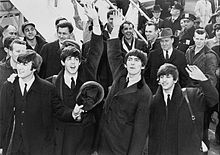 Black-and-white picture of four young men outdoors in front of a staircase, surrounded by a large assembled crowd. All four are waving to the crowd.