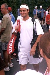 A dark-haired man in all white clothing, and caring a redish-black bag on his right shoulder and a black one on the left shoulder