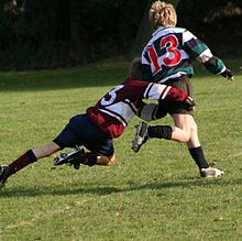 A child running away from camera in green and black hooped rugby jersey is in the process of being tackled around the hips and legs by an opponent.