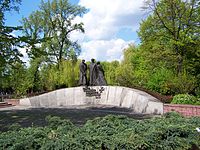 A low stone wall, curving upward. Three statues of Polish soldiers are mounted at its center. Below the statues, Text is mounted as per caption.