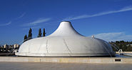 A gray semi-hemispherical structure with a protruding top, standing in the middle of a fountain, with several jets of water falling on the structure