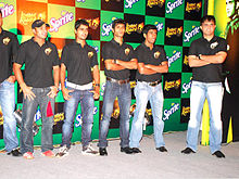 A group of men standing, wearing black t-shirts and blue faded jeans. All of them are looking to the left of the image. The backdrop has alternate black and green boxes.