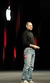 Full-length portrait of man about fifty wearing jeans and a black turtleneck shirt, standing in front of a dark curtain with a white Apple logo