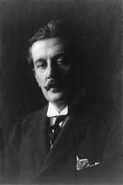 Head and shoulders portrait in heavy shadow, depicting a dark-haired man with a curling moustache, wearing a winged collar with a tie and with a handkerchief protruding from the top pocket